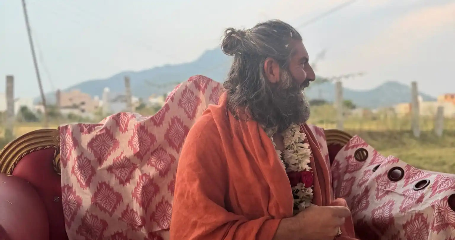 Bearded person wearing an orange robe seated on a patterned couch.