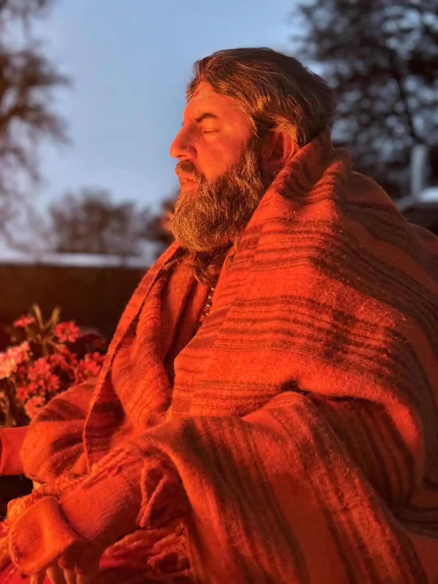 A bearded person wrapped in a red blanket sits with eyes closed in warm lighting.