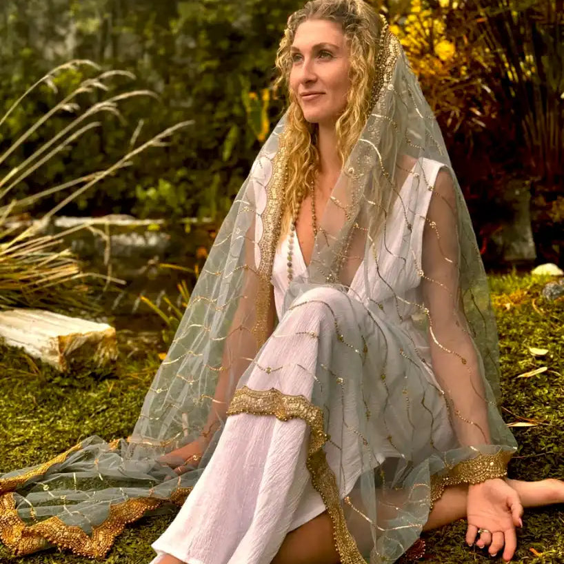 A bride wearing a flowing lace veil and bohemian-style wedding dress sits barefoot on grass.