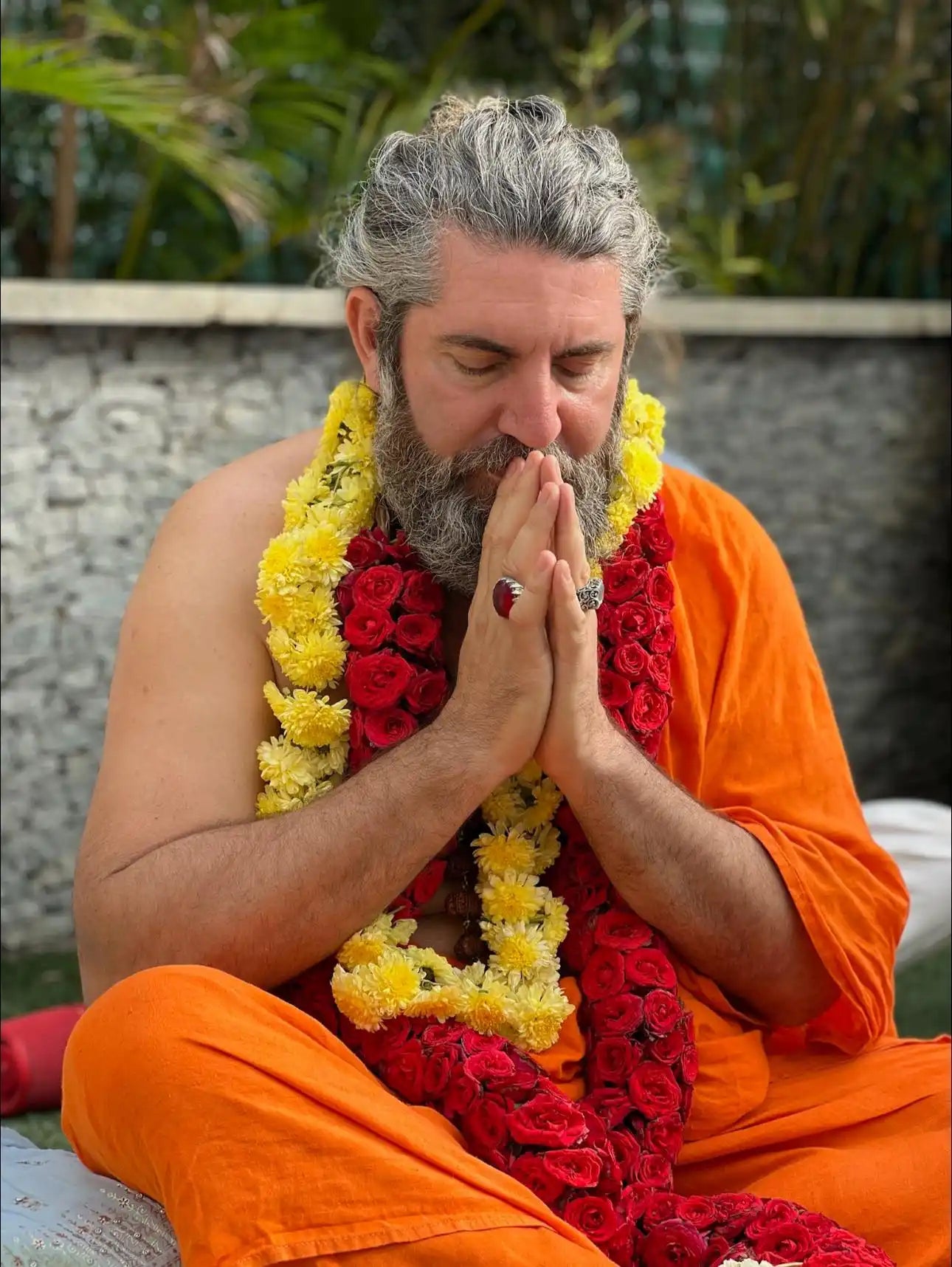 A person in orange robes and red-yellow flower garlands meditating with clasped hands.