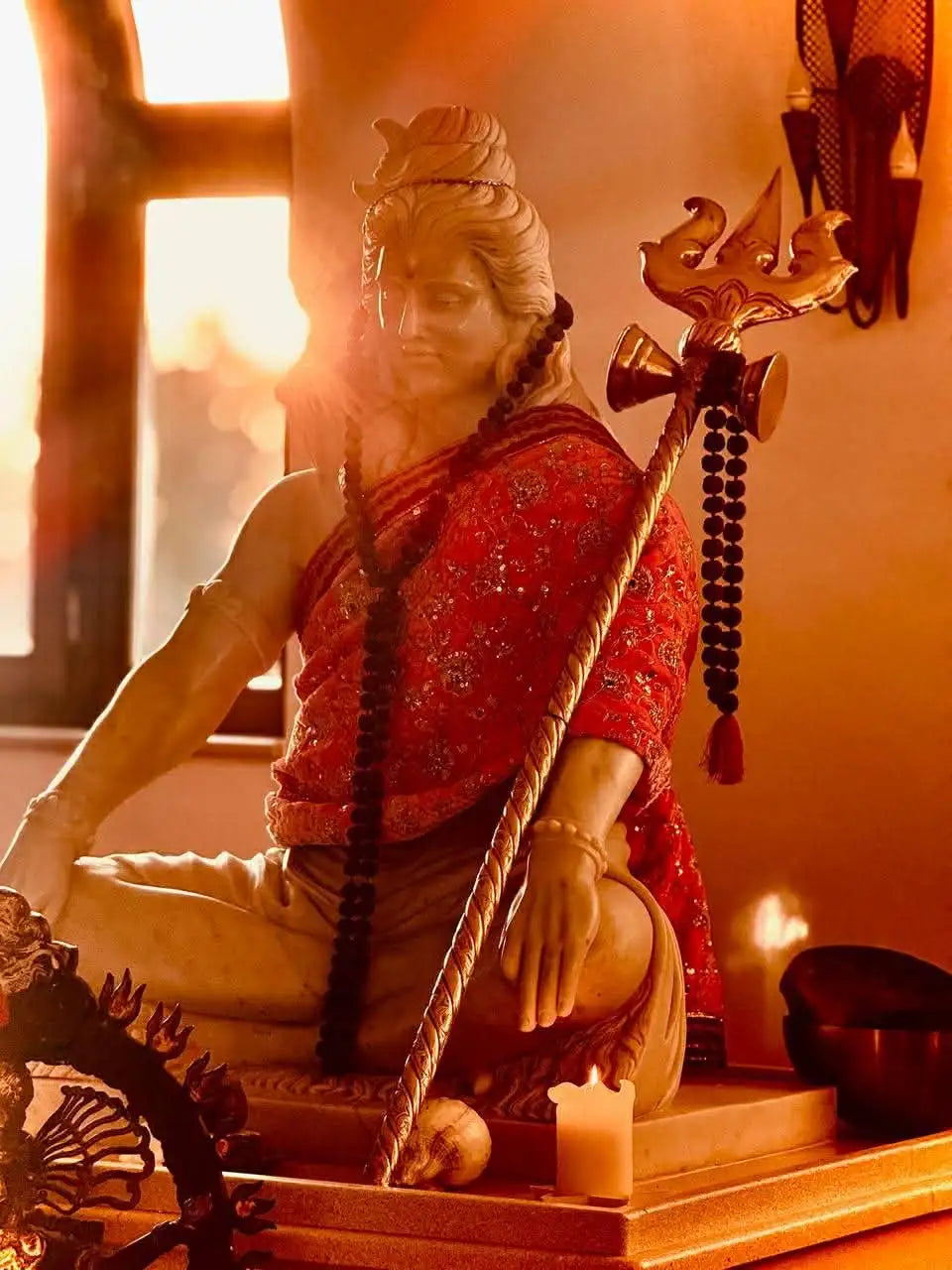 A detailed, golden-hued statue of a seated deity in traditional red and gold attire holding a trident.