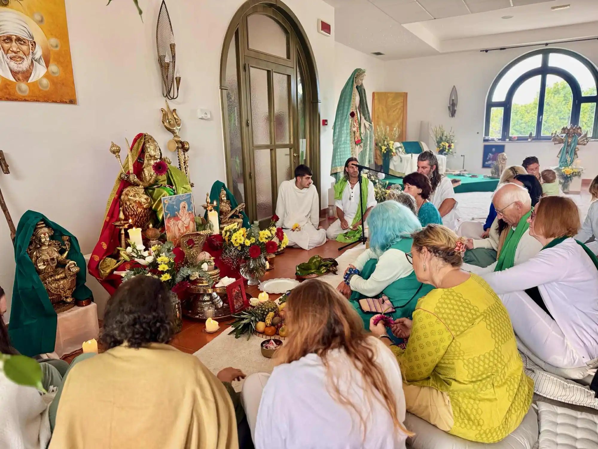 A diverse group of people sitting on the floor in a spiritual gathering, surrounded by religious altars and decorations.