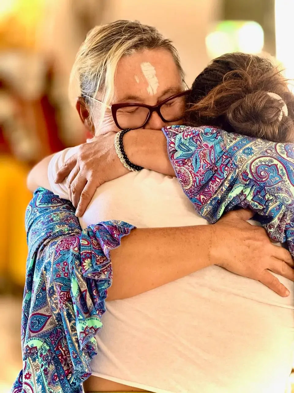 A woman in a vibrant blue, purple, and green paisley-patterned blouse with ruffled sleeves embraces another person.
