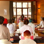 A person with long hair, wearing a white robe and sitting cross-legged on a mat in front of an audience.