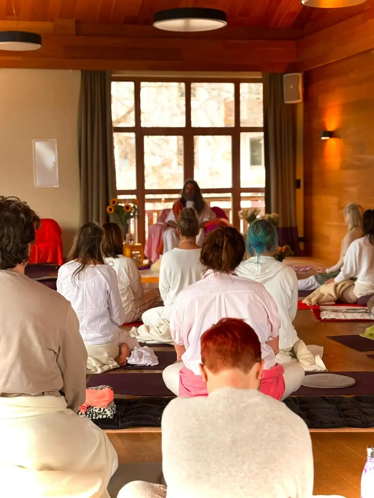 A person with long hair, wearing a white robe and sitting cross-legged on a mat in front of an audience.