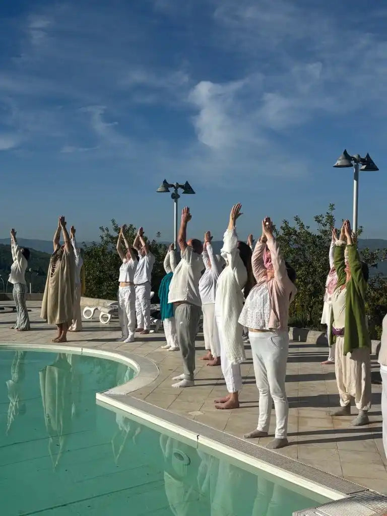 A group of people in light-colored clothing performing a yoga pose outdoors by a pool.