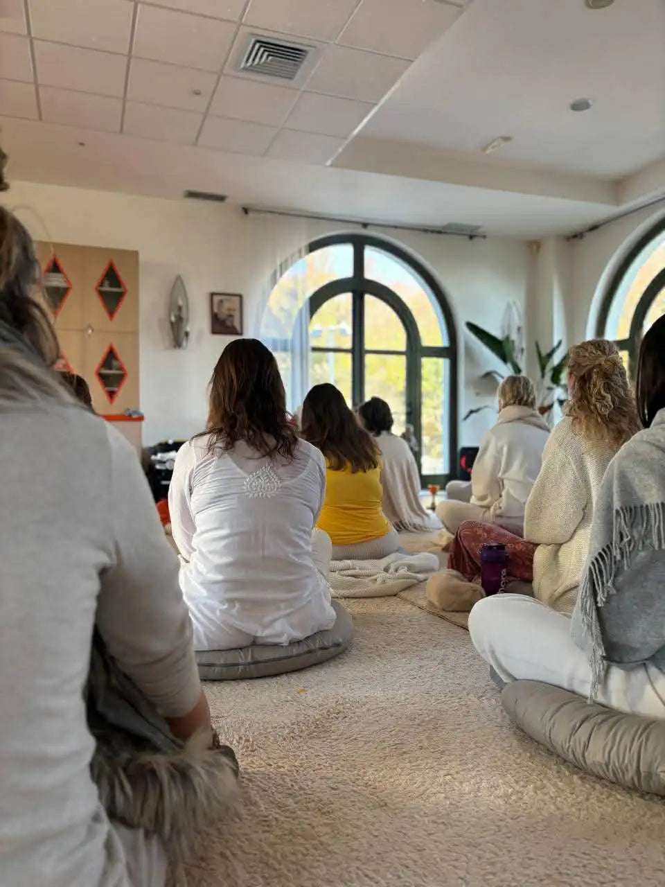 A group of people seated cross-legged on soft, cushioned mats in a bright, airy room with arched windows.