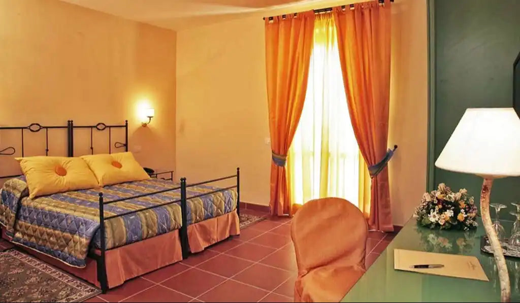 A hotel room with a wrought-iron bed frame, patterned bedspread, and a desk with a lamp and flowers.