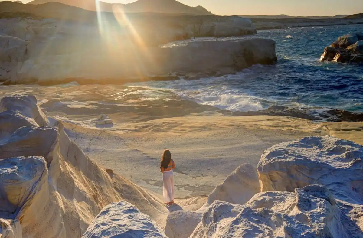 A lone woman standing on a rocky, white-colored beach at sunset.