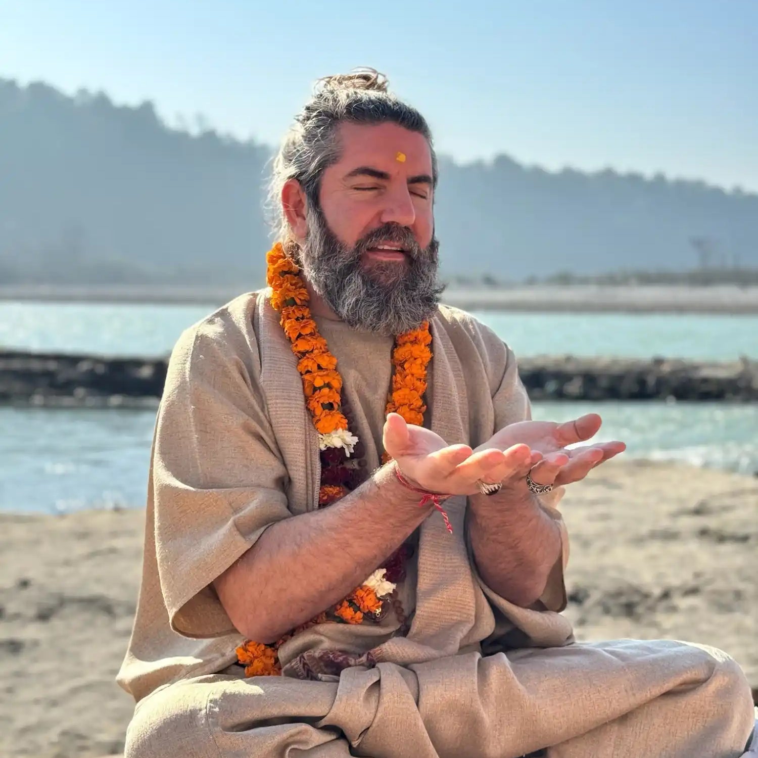 A man with a full beard and hair tied in a bun, wearing a traditional beige robe and an orange marigold garland, sitting cross-legged with hands open in a meditative pose.