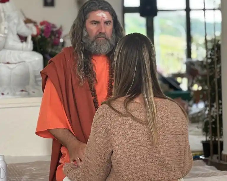 A man with a beard and long hair wearing orange robes, facing a woman with long hair in a brown sweater and white pants, both seated on a mat.