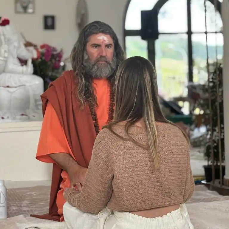 A man with a beard and long hair wearing orange robes, facing a woman with long hair in a brown sweater and white pants, both seated on a mat.