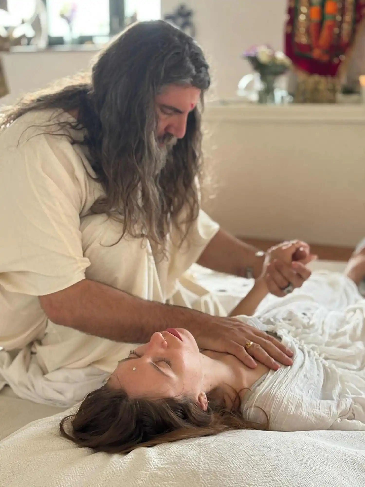 A man in a light-colored robe with long hair and a beard, performing a healing or therapeutic touch on a woman lying down.