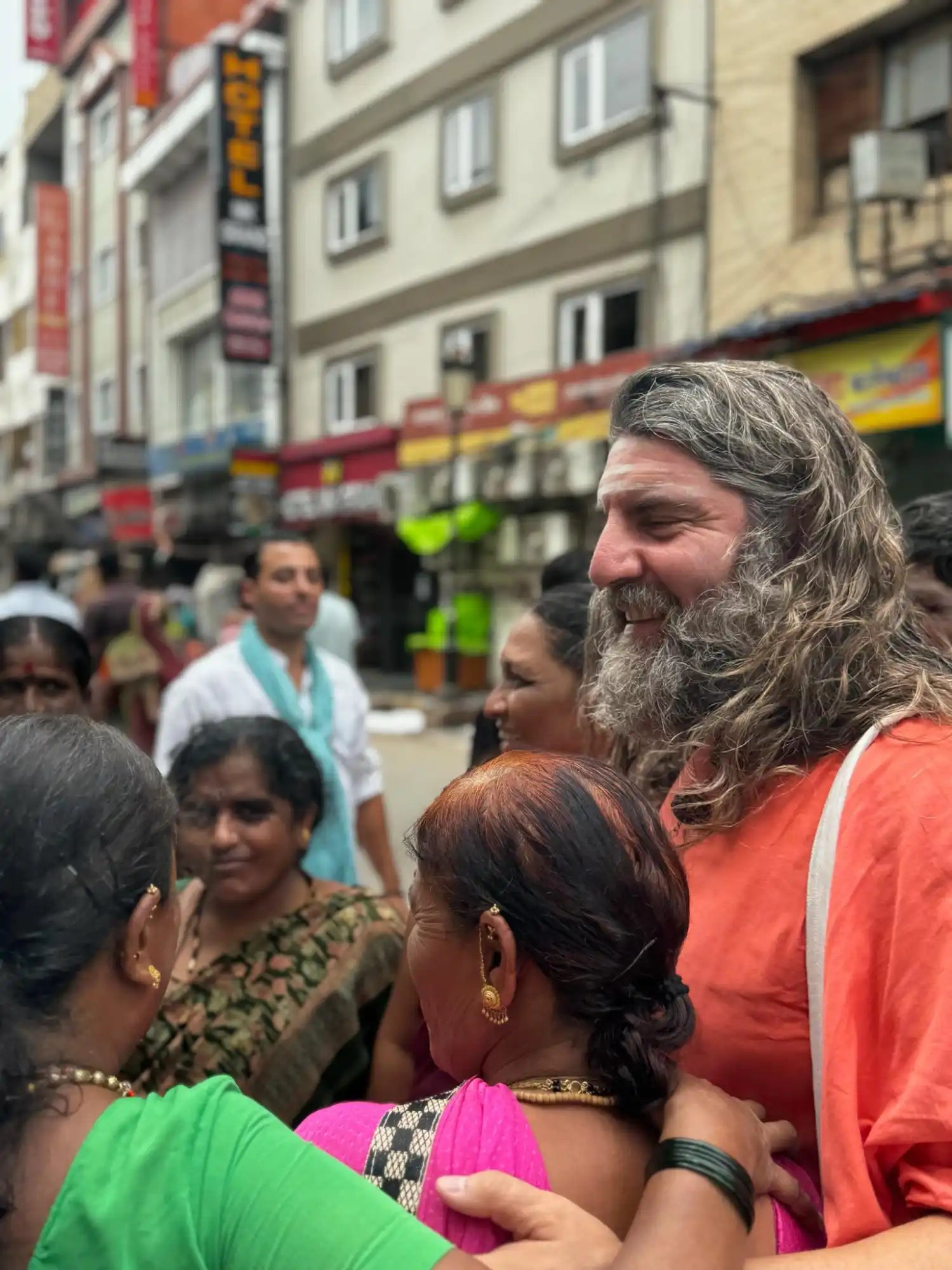 A man with a long beard and hair, wearing an orange robe, interacting with people in a busy street.
