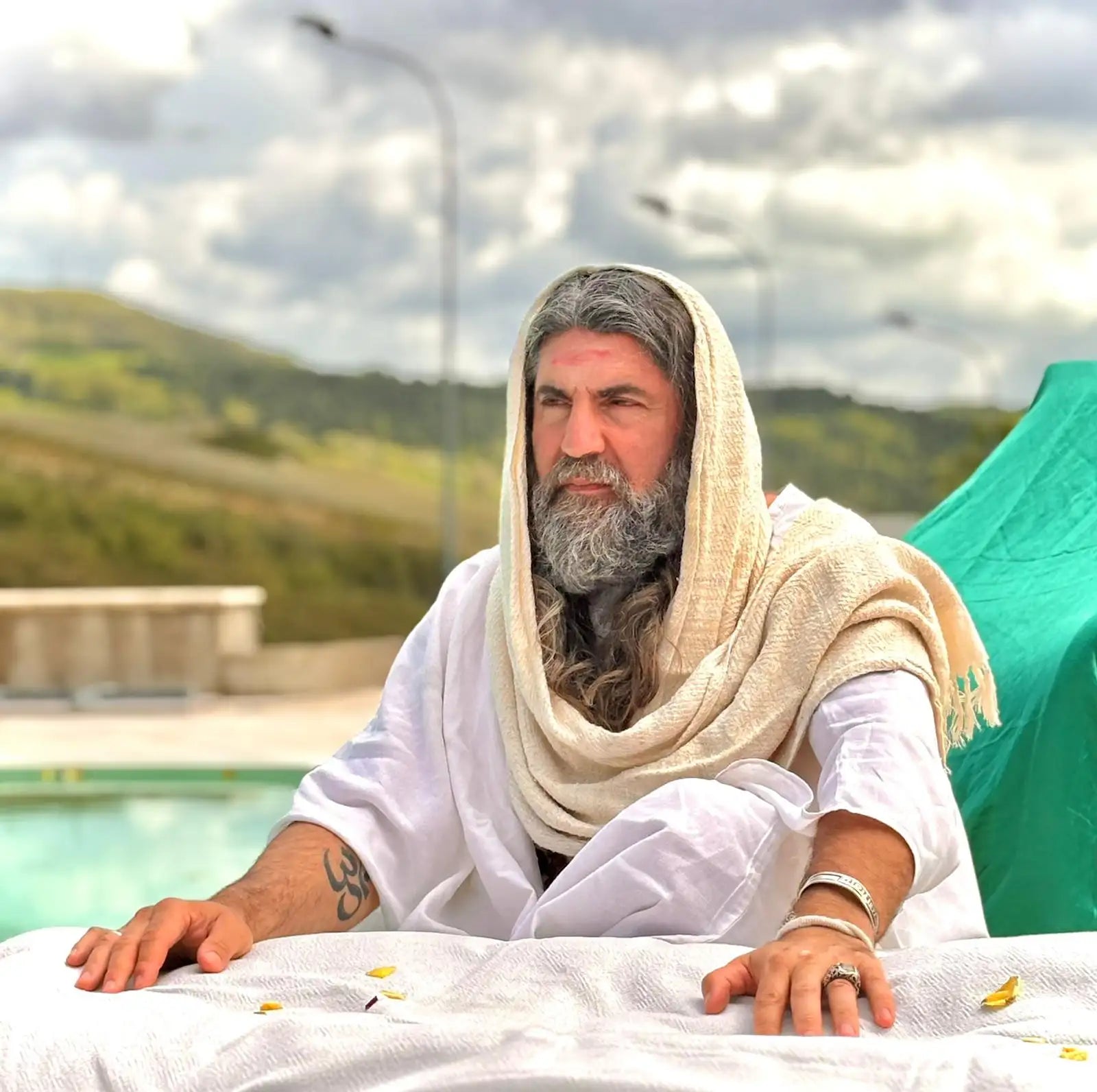 A man with long gray hair and a full beard, wearing an off-white draped robe over white clothing.