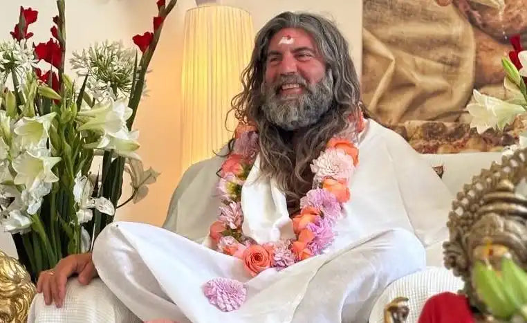 A smiling man with long gray hair and beard, wearing a white robe and a floral garland.
