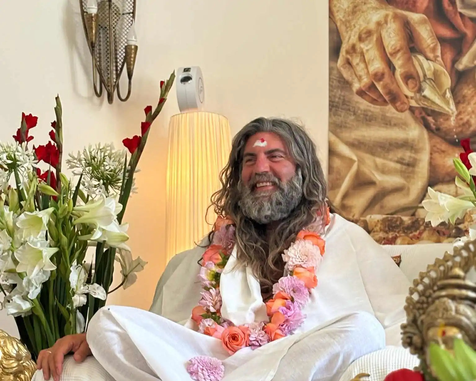 A smiling man with long gray hair and a beard, wearing a white robe and floral garlands.