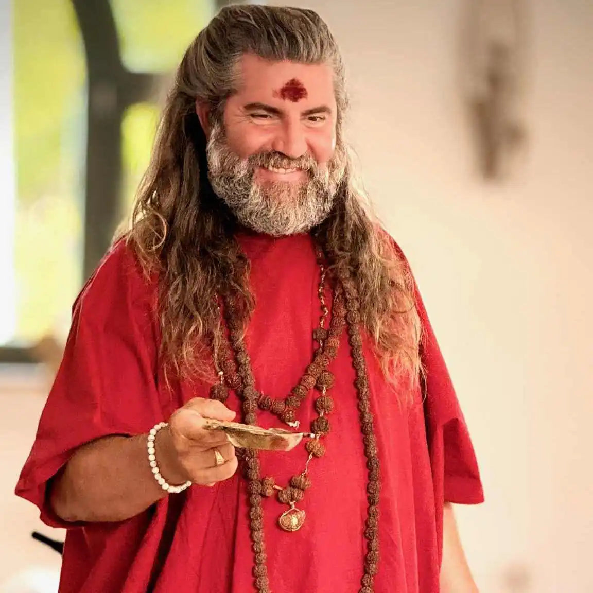 Smiling man with long hair and beard, wearing a red robe and beaded necklaces, holding a small brass bowl.