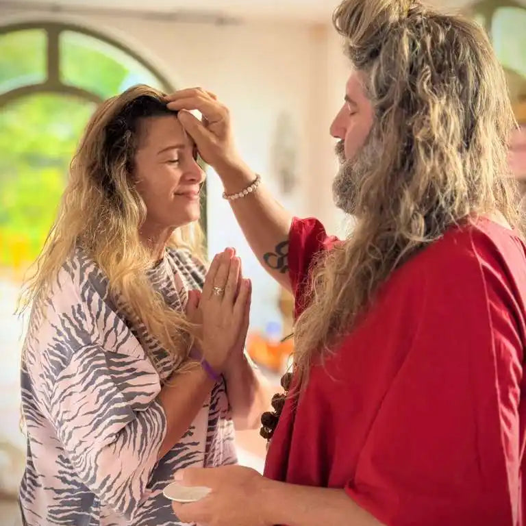 A man with long hair and beard in red robes performing a blessing on a woman with long wavy hair.