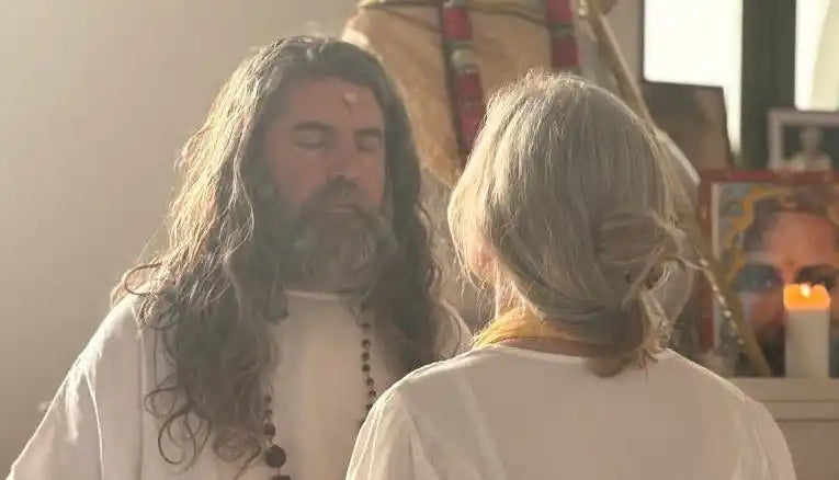 A man with long hair and a beard, wearing a white robe and prayer beads, with eyes closed in meditation.