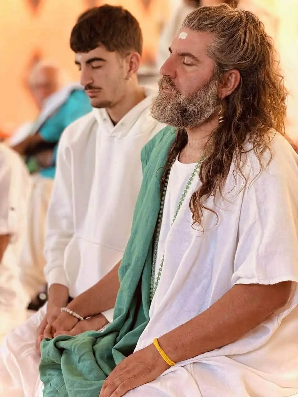 A man with long, wavy brown hair and a full beard wearing white robes draped over his shoulders.