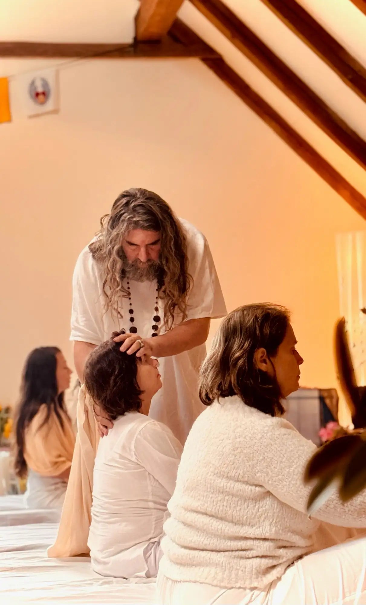 A man with long, wavy gray hair and a beard wears a simple white t-shirt while holding a dark beaded necklace.