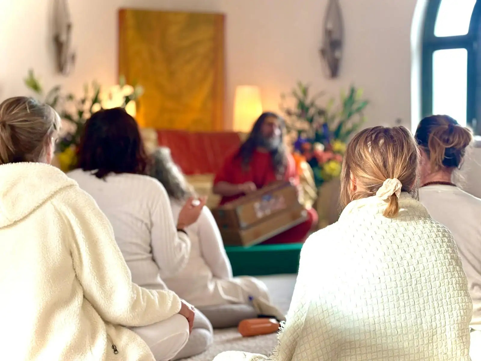 A person with long hair and a beard, wearing a red robe, sits in the center of a room playing a wooden instrument.