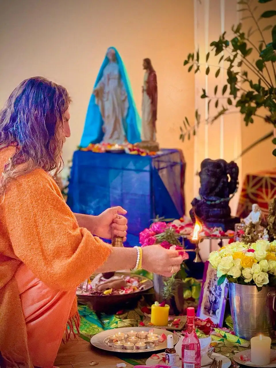 A person in an orange robe lights a candle at a vibrant altar adorned with flowers, statues, and offerings.