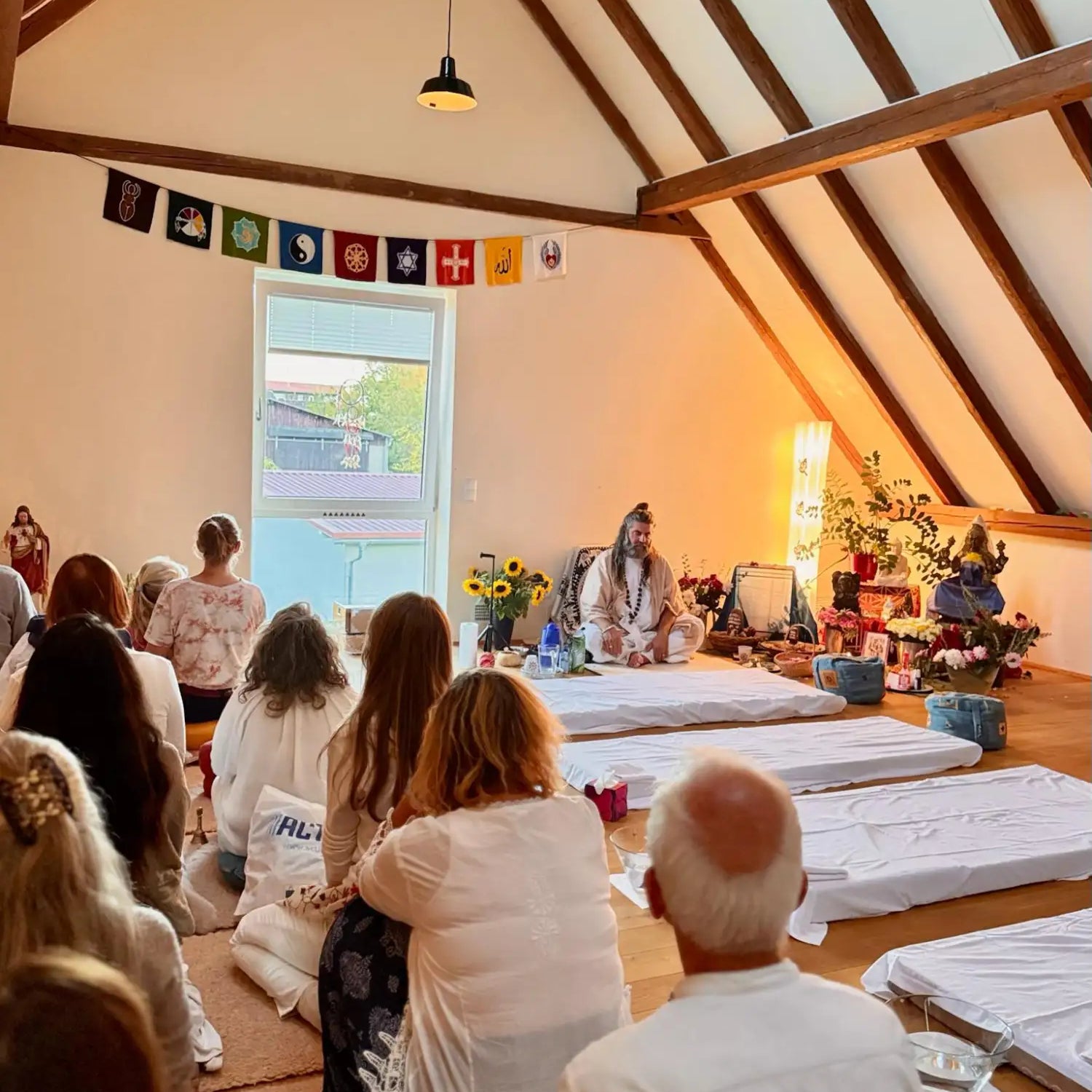 A serene meditation space with wooden beams, string lights, and a central figure seated on a mat surrounded by attendees in white clothing.