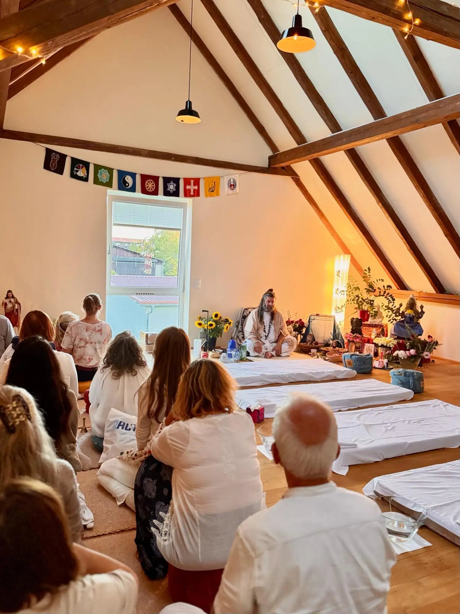 A serene meditation space with wooden beams, string lights, and a central figure seated on a mat surrounded by attendees in white clothing.