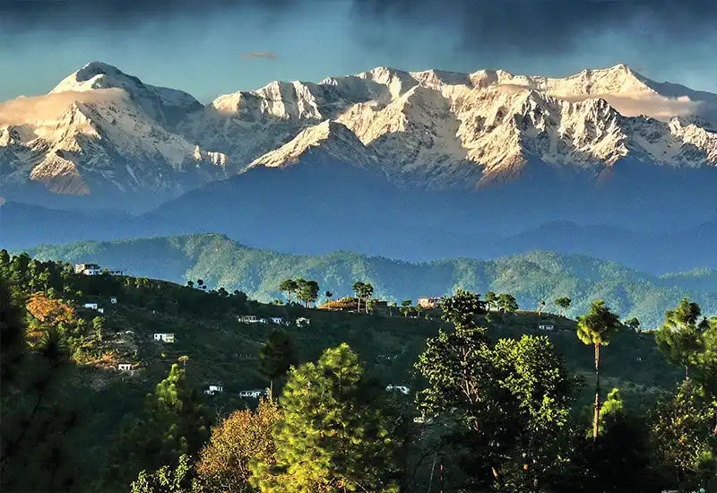 A serene mountain landscape featuring snow-capped peaks, verdant hills dotted with houses, and coniferous trees in the foreground.