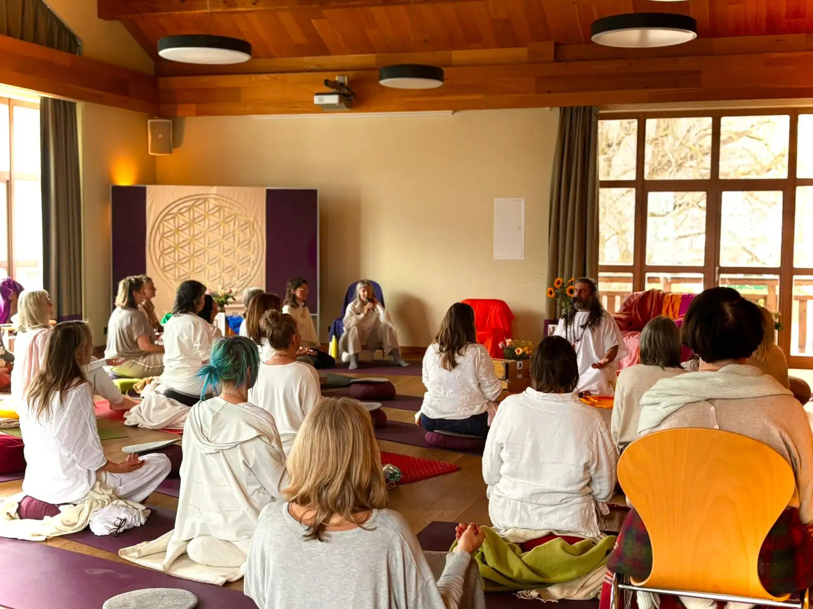 A serene yoga studio with wooden beams, large windows letting in natural light, and a group of people seated on mats facing an instructor who is sitting cross-legged at the front.