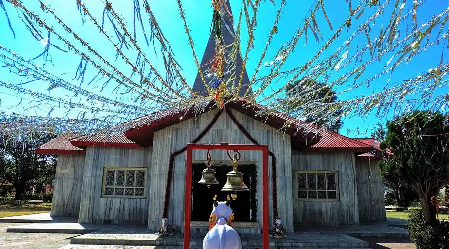 A temple with a pointed roof and walls made of light-colored, possibly wooden, planks, decorated with numerous festive streamers and two large bells hanging at the entrance.