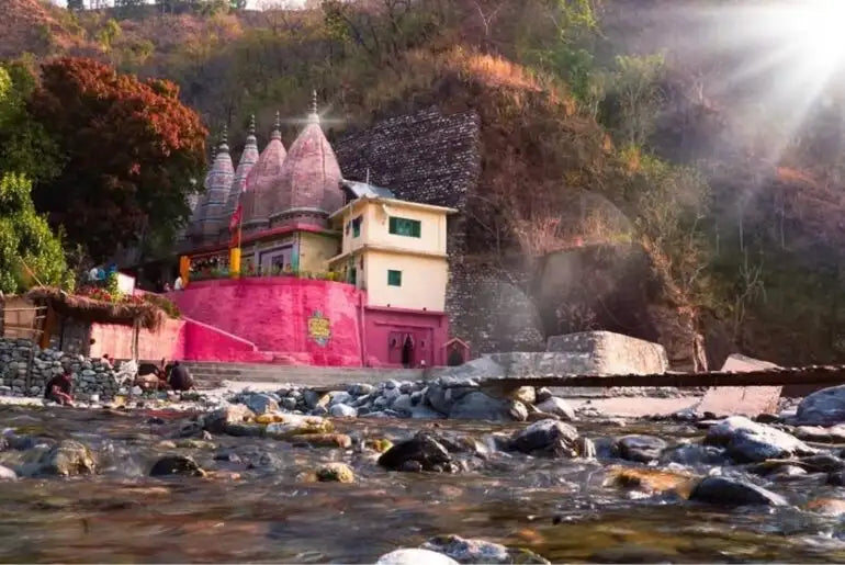 A vibrant pink temple with multiple conical spires and a cream-colored annex stands beside a rocky riverbank.