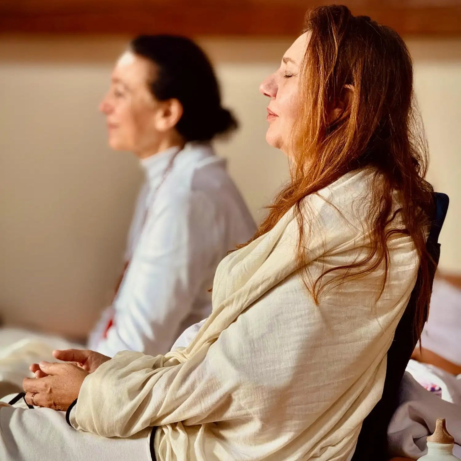 A woman in a cream-colored, long-sleeved robe with black trim sits cross-legged in meditation.