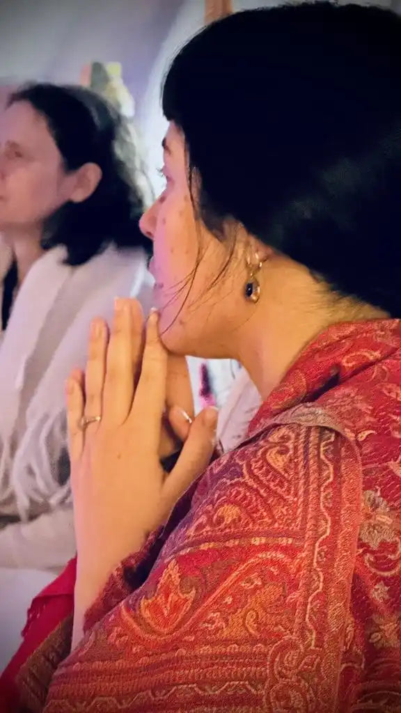 A woman with dark hair and bangs, wearing a red patterned shawl, with her hands clasped in front of her.