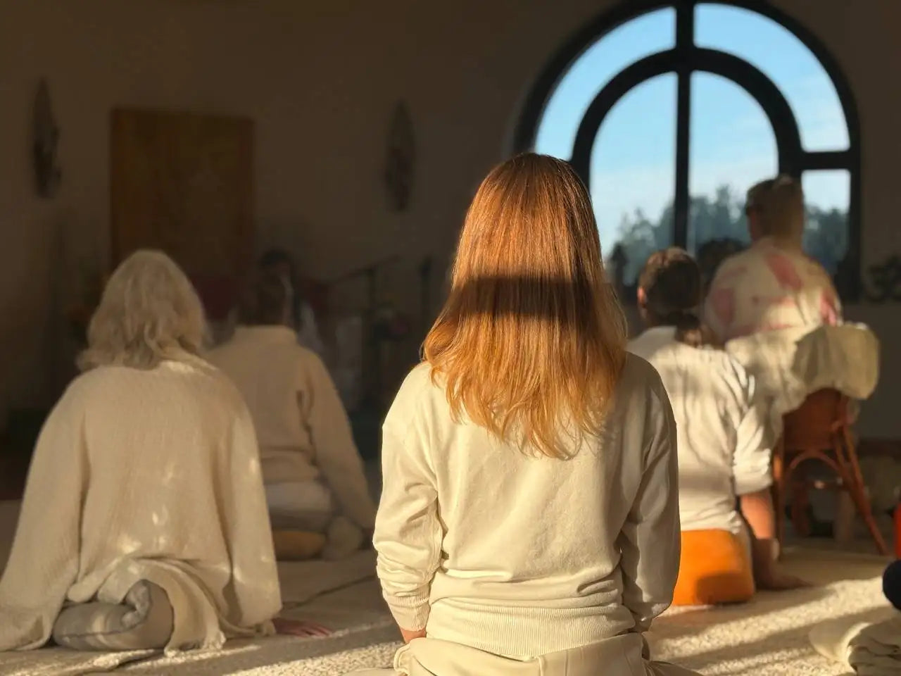 A woman with long, straight auburn hair sits cross-legged in front of an arched window.