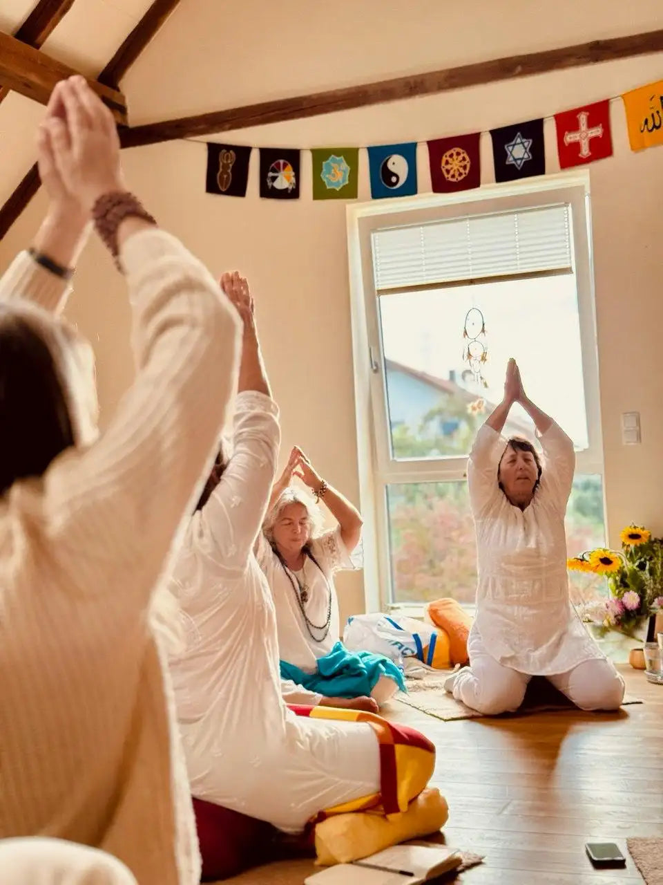 A woman in a white long-sleeved top and pants kneels on the floor with her hands pressed together above her head, wearing multiple bracelets.